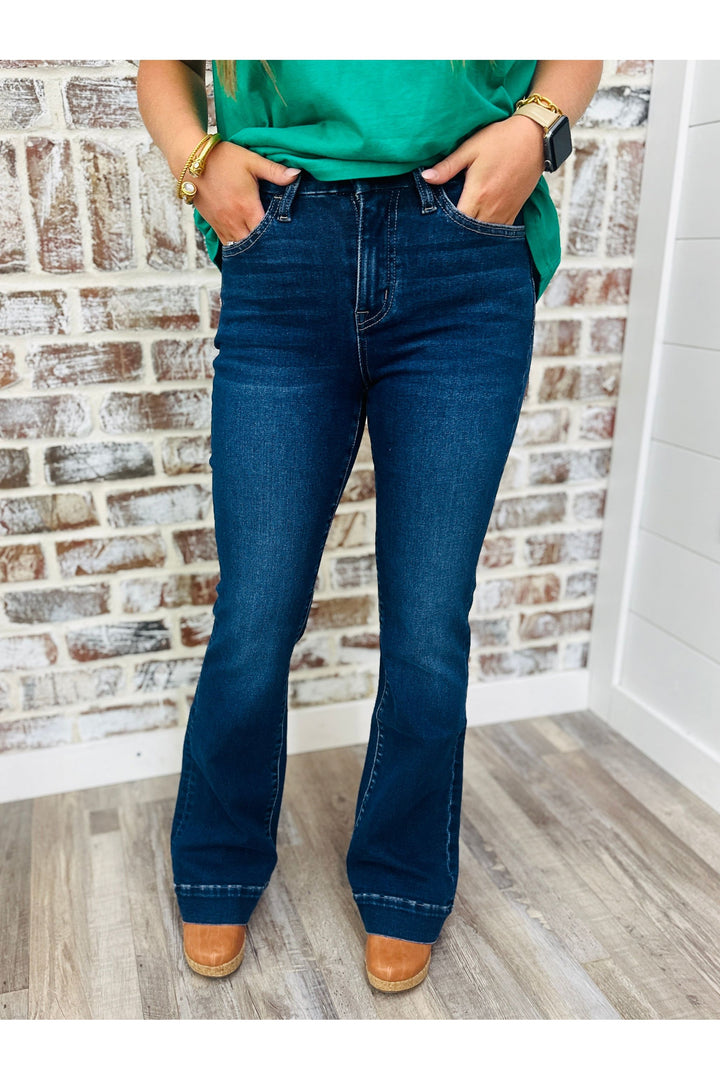 Woman wearing dark denim flare jeans standing against a brick wall.
