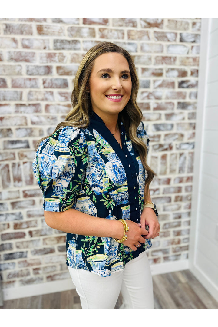 Woman wearing patterned blouse and white pants standing against a brick wall.