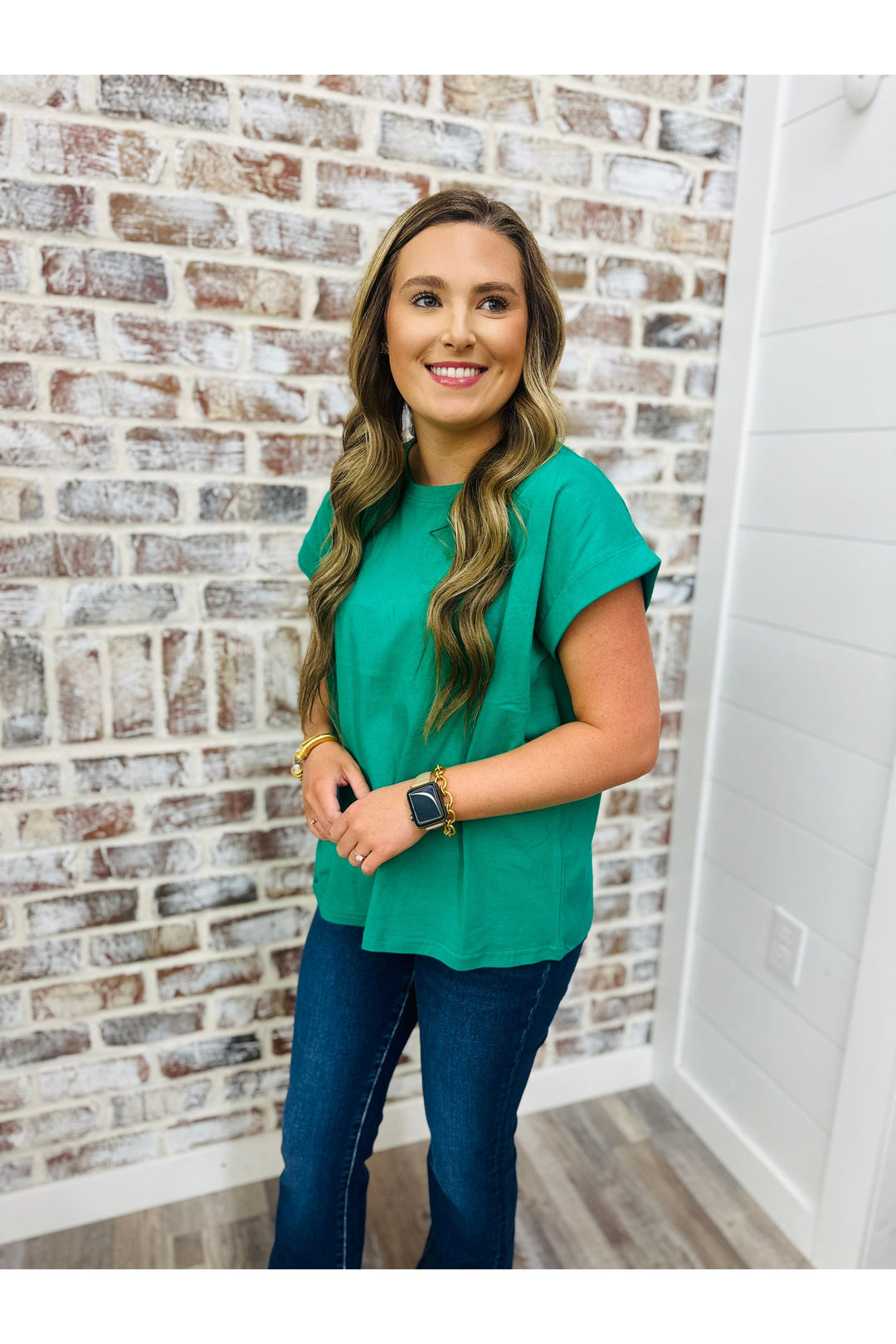 Woman wearing emerald green cotton shirt with jeans standing against a brick wall.