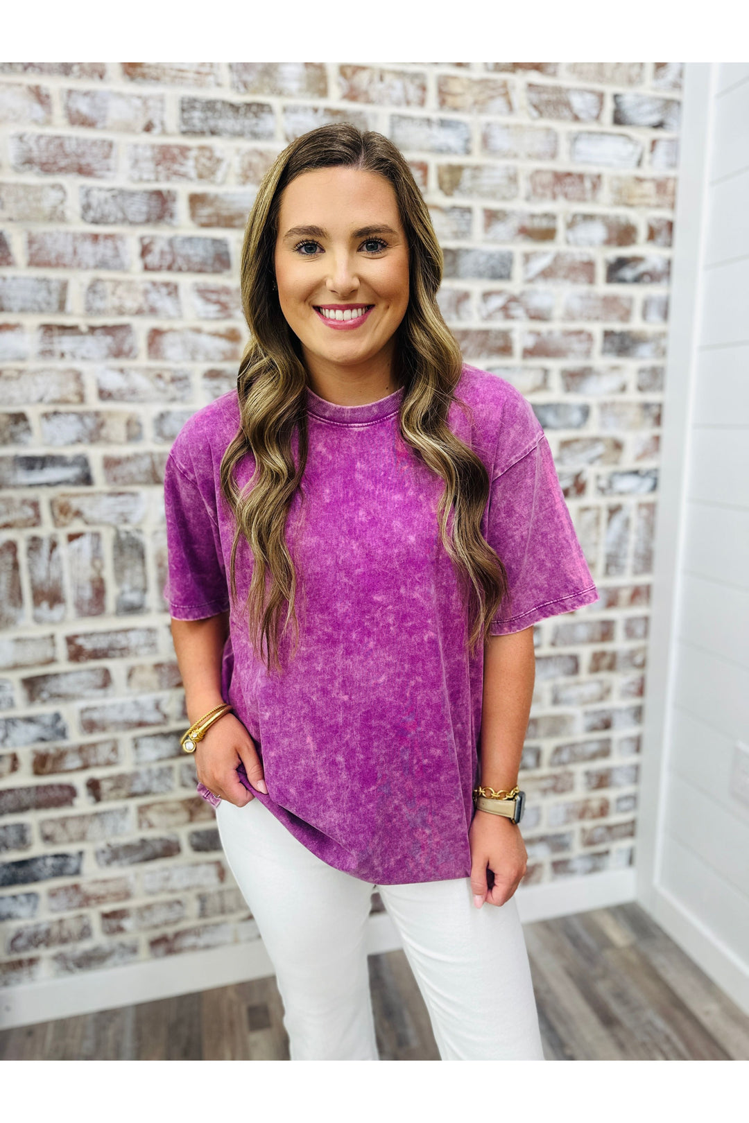 Woman wearing oversized plum acid washed tshirt with white jeans standing against a brick wall.