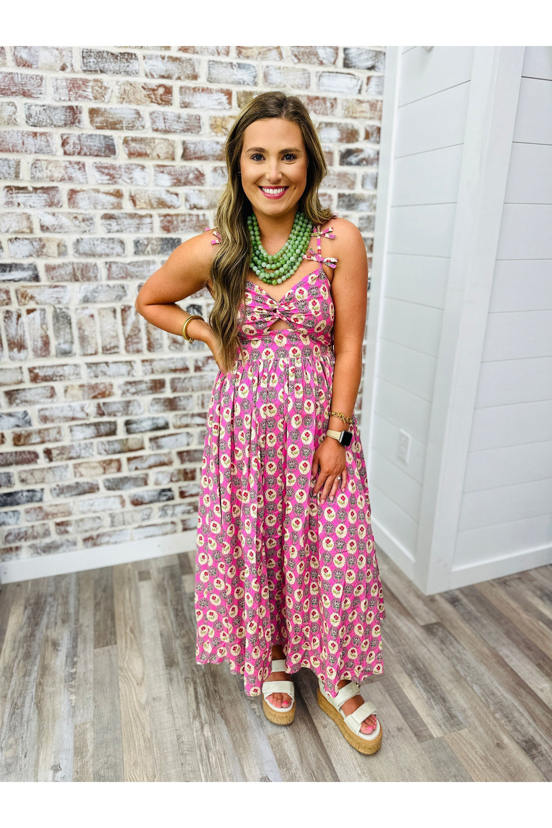 Woman wearing a pink floral dress with front cut out and bow detail on the strap. Paired with a olive beaded necklace. 