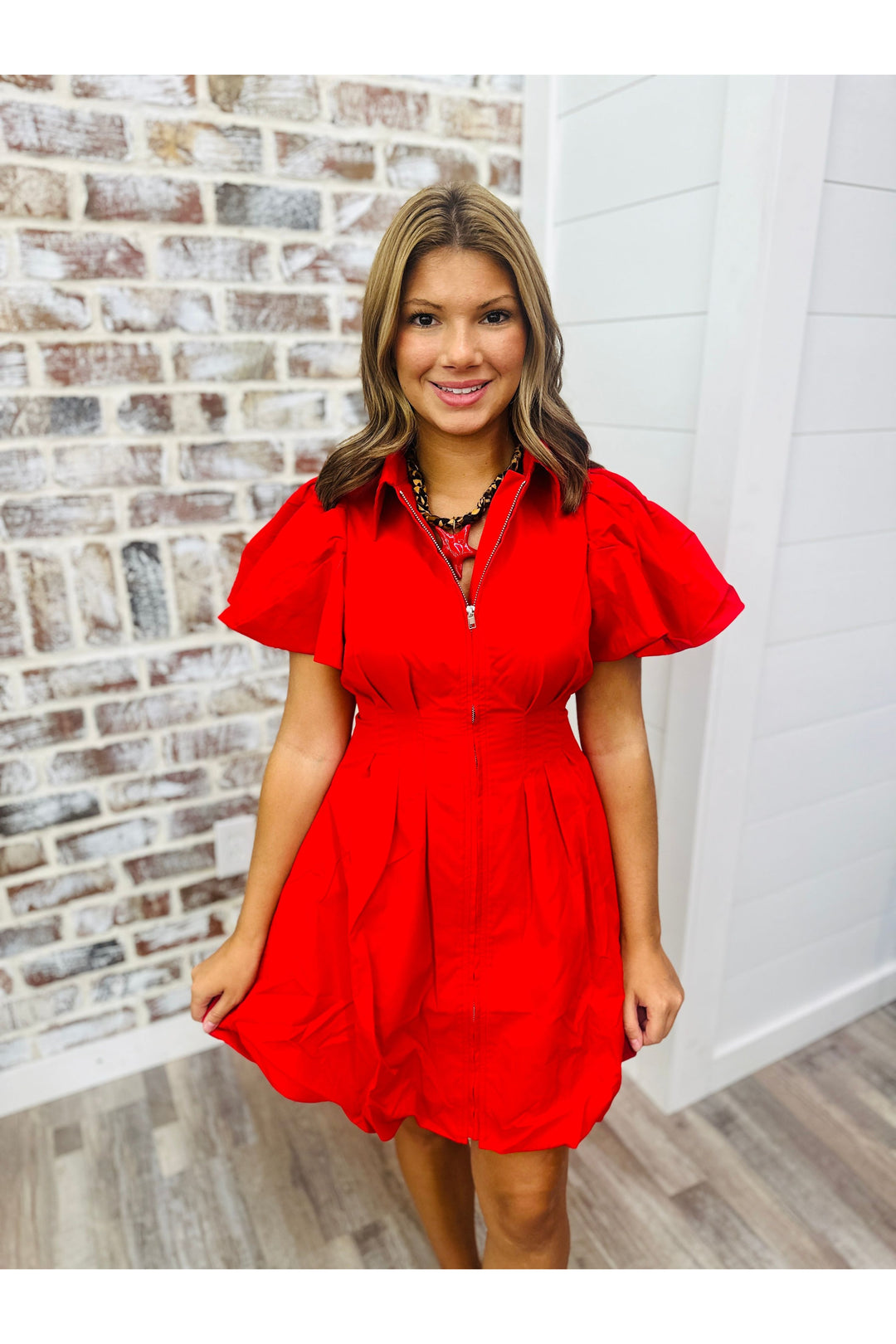 Women wearing a red dress with a cinched waist standing against a brick wall. Paired with a leopard scarf necklace with a red star.