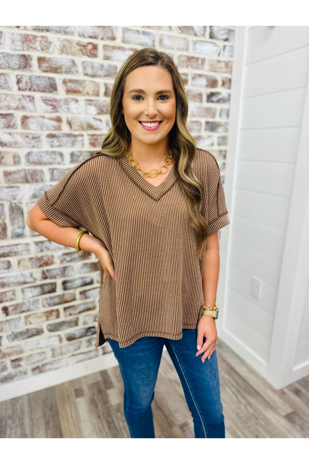 Woman wearing a brown short sleeve waffle top with jeans standing against a brick wall.