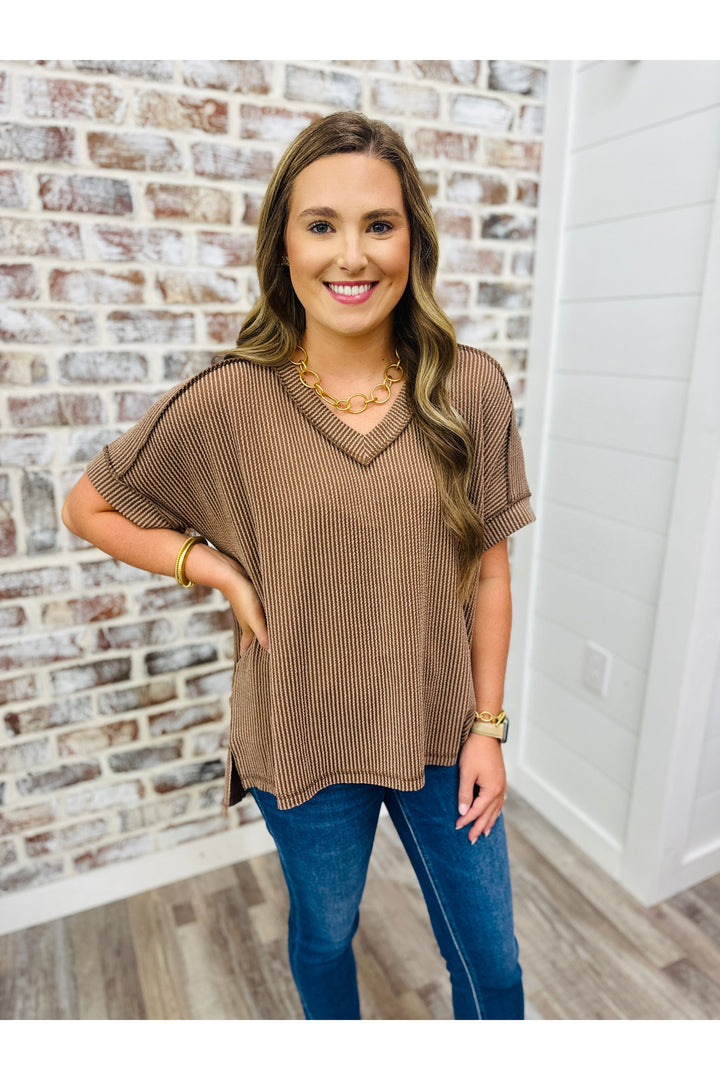 Woman wearing a brown short sleeve waffle top with jeans standing against a brick wall.