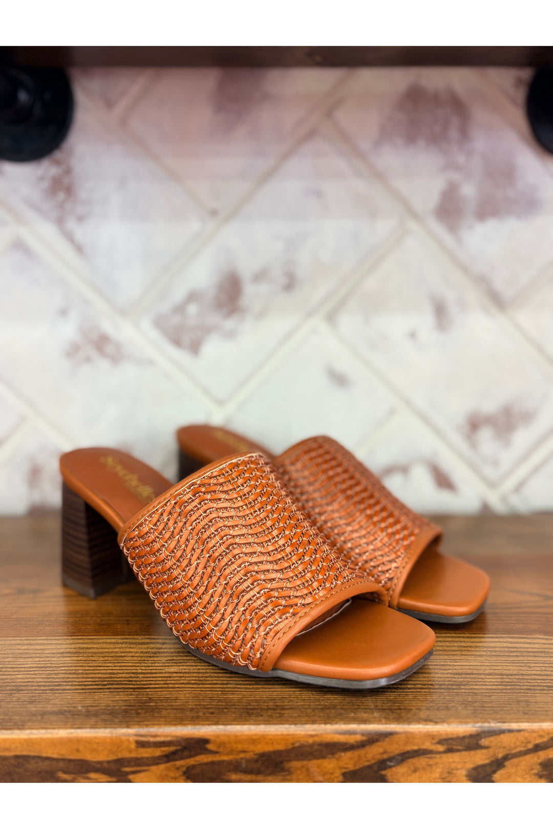 Brown woven slide sandals on a wooden surface with a patterned wall in the background