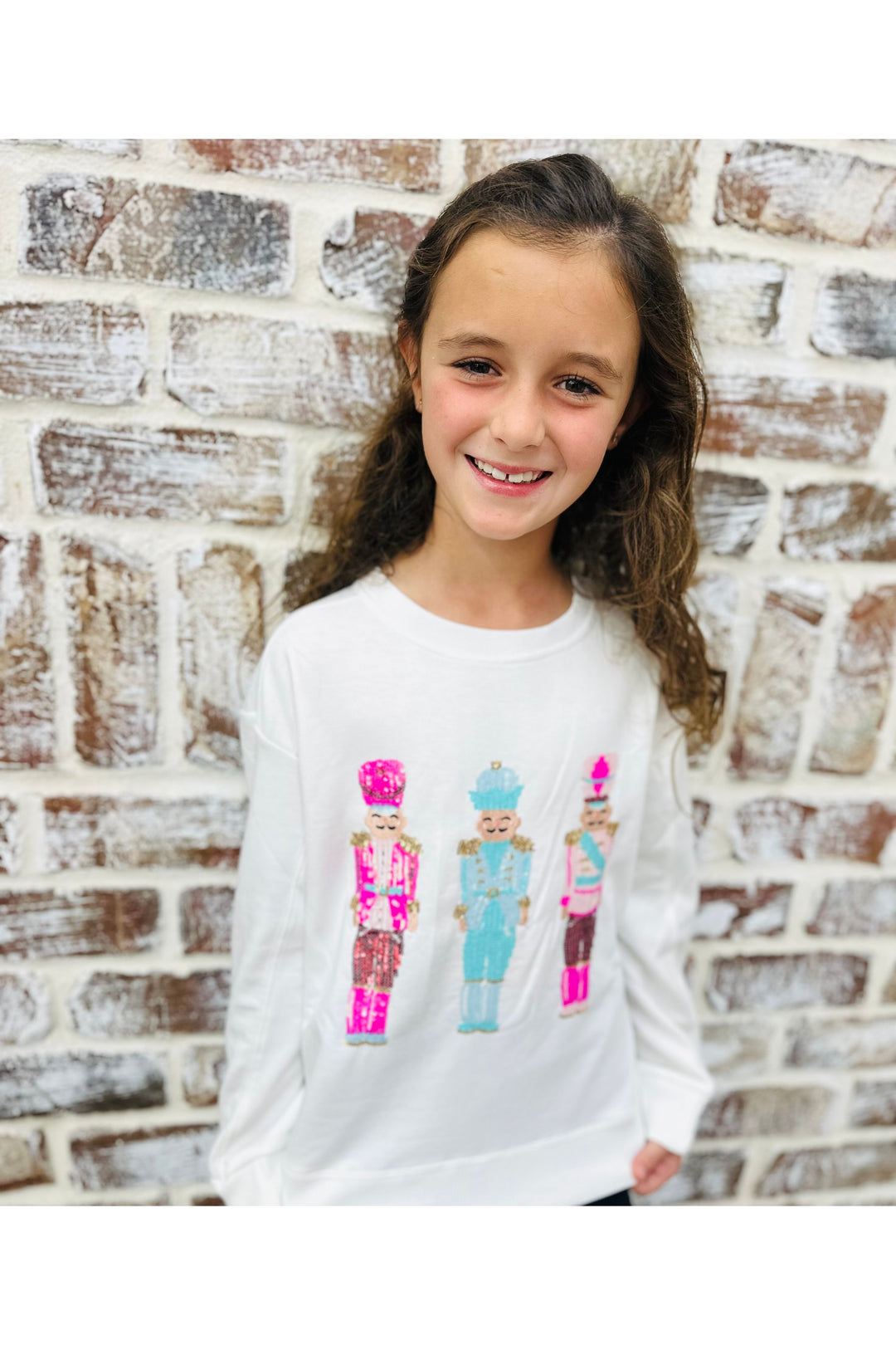 Young girl wearing a white sweatshirt with colorful nutcracker design against a brick wall.