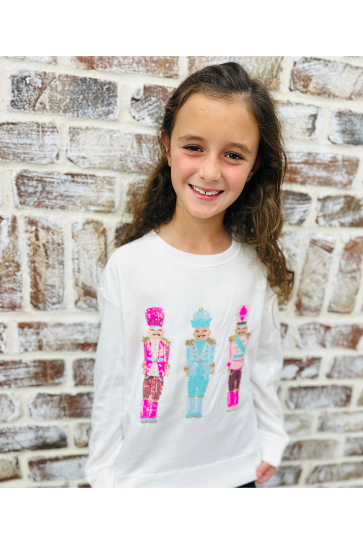 Young girl wearing a white sweatshirt with colorful nutcracker design against a brick wall.