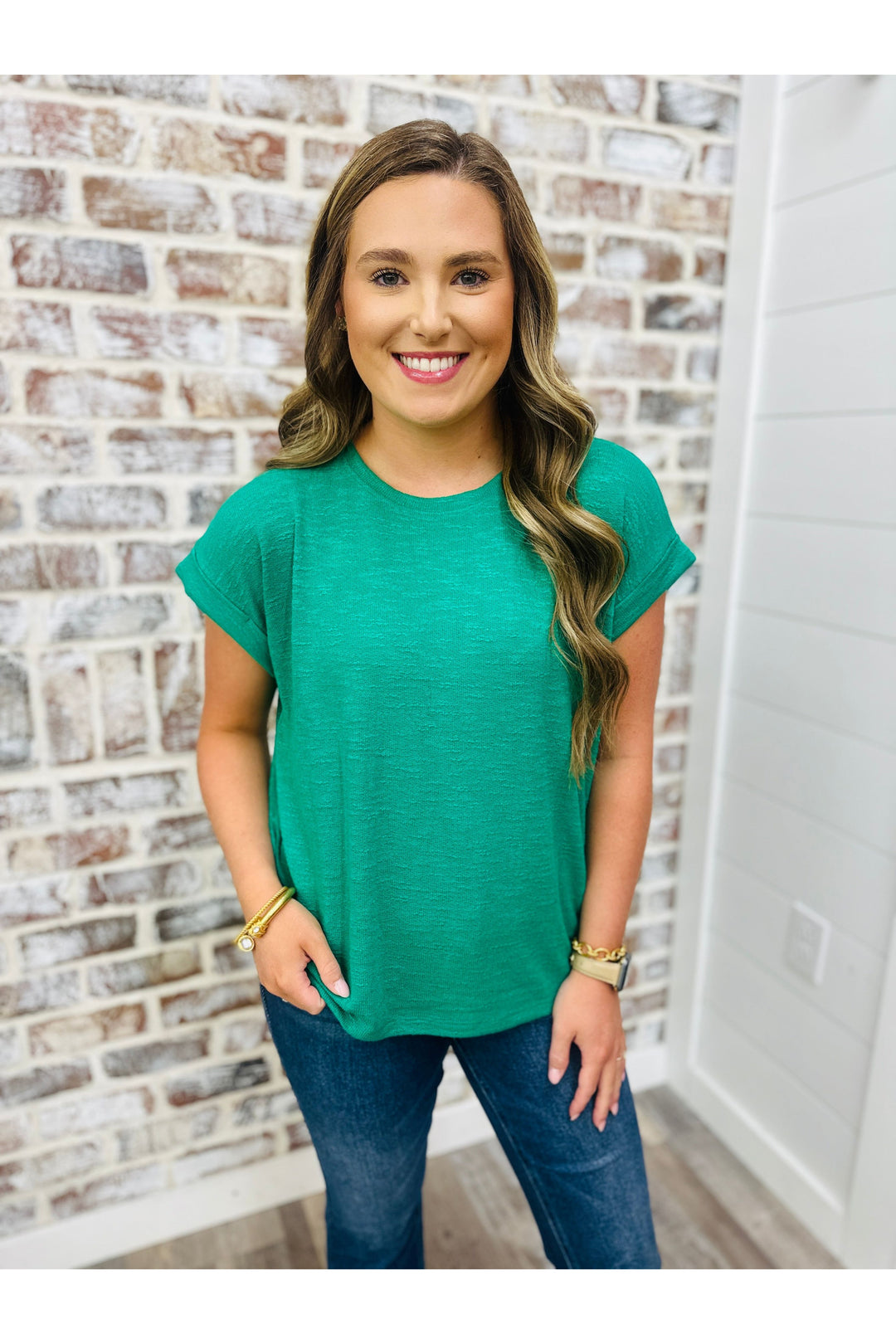 Woman wearing emerald green solid knit top with jeans standing against a brick wall.