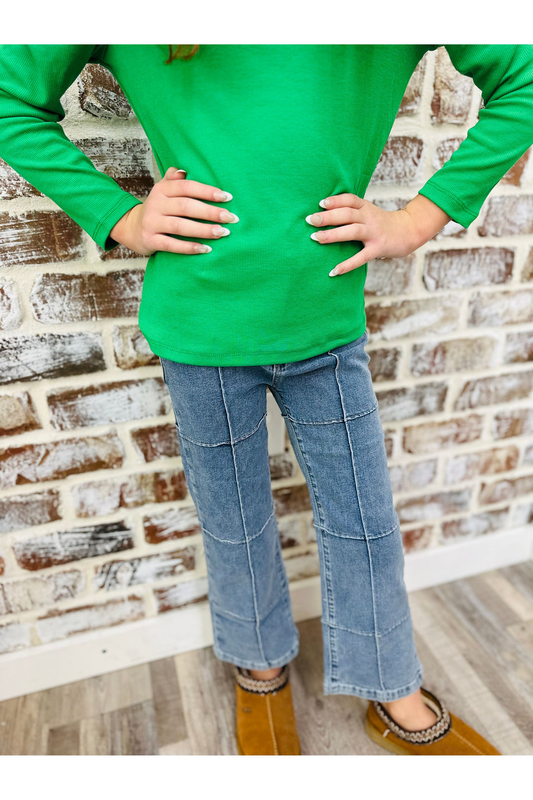 Person wearing a green long-sleeve shirt and blue jeans standing against a brick wall.