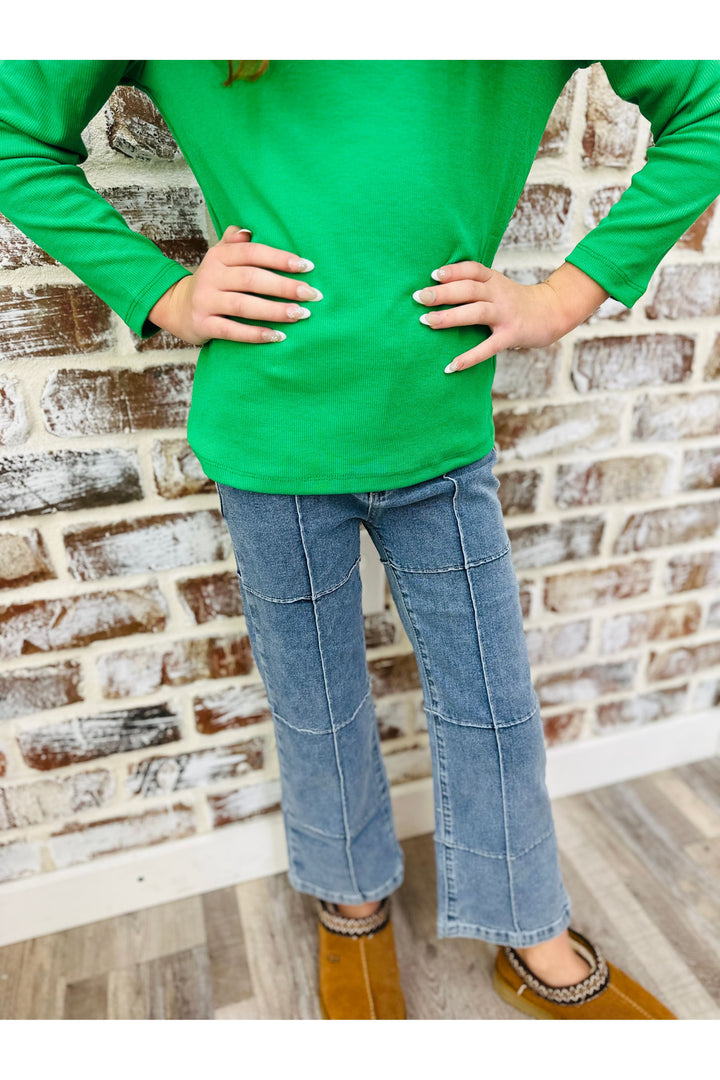 Person wearing a green long-sleeve shirt and blue jeans standing against a brick wall.