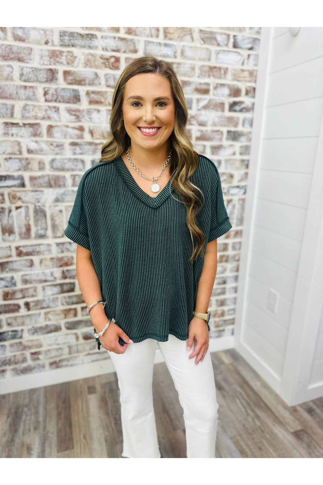 Woman wearing a hunter green waffle top paired with white pants standing against a brick wall.