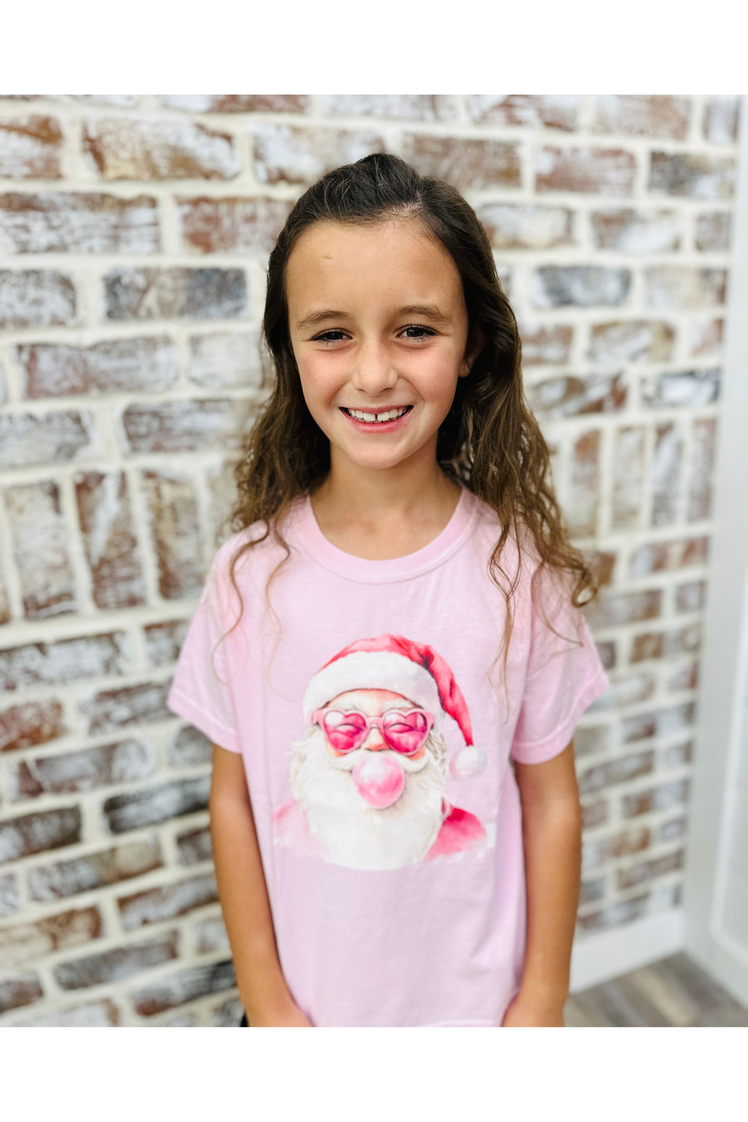 Young girl wearing a pink t-shirt with a Santa Claus graphic against a brick wall.