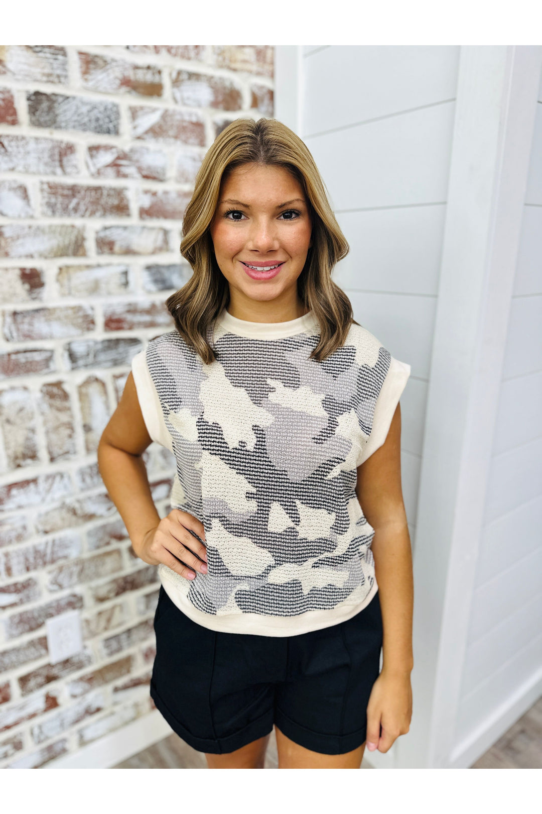Woman wearing camoflauge knit top with black shorts standing against a brick wall.