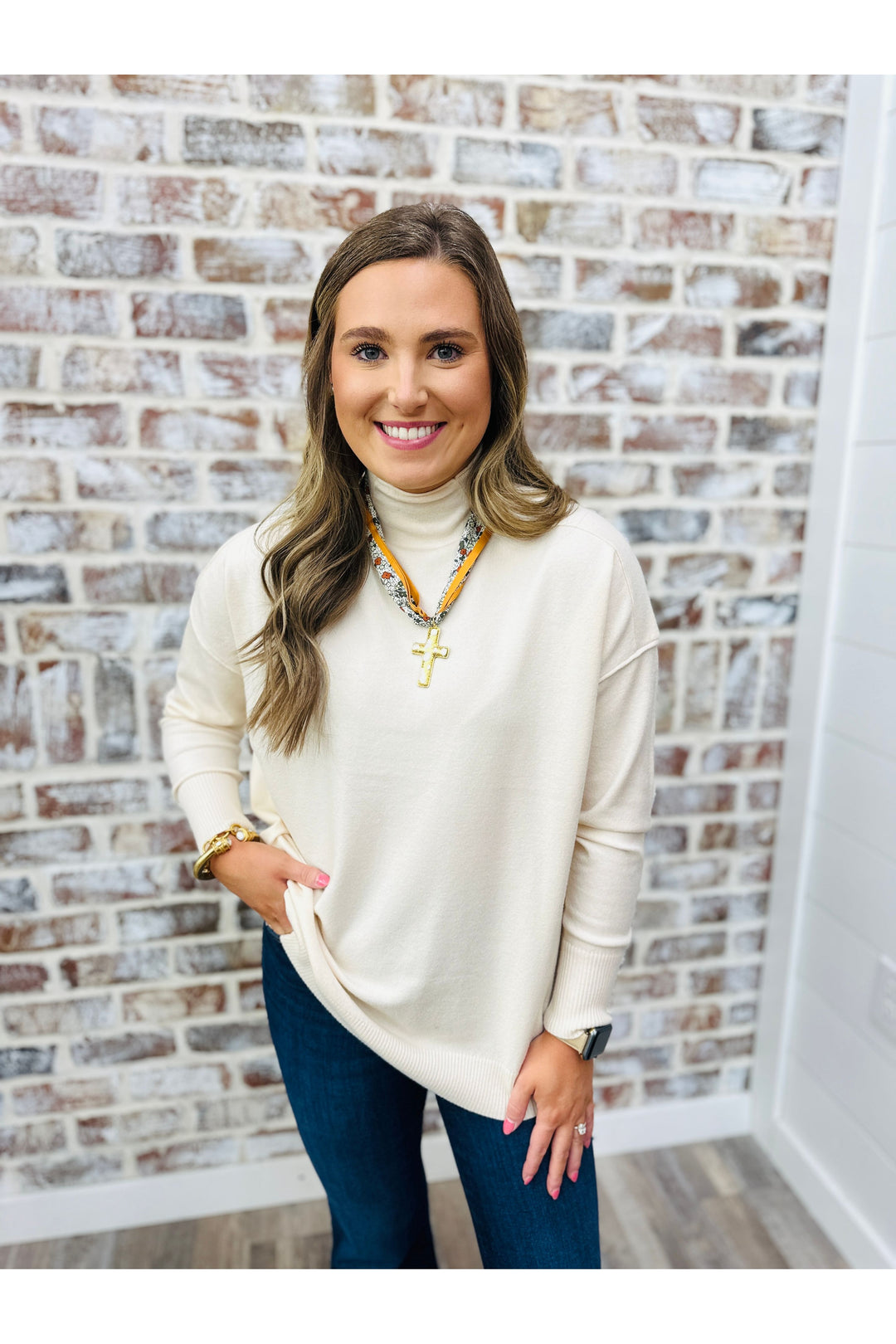 Woman wearing a cream sweater and blue jeans standing against a brick wall.