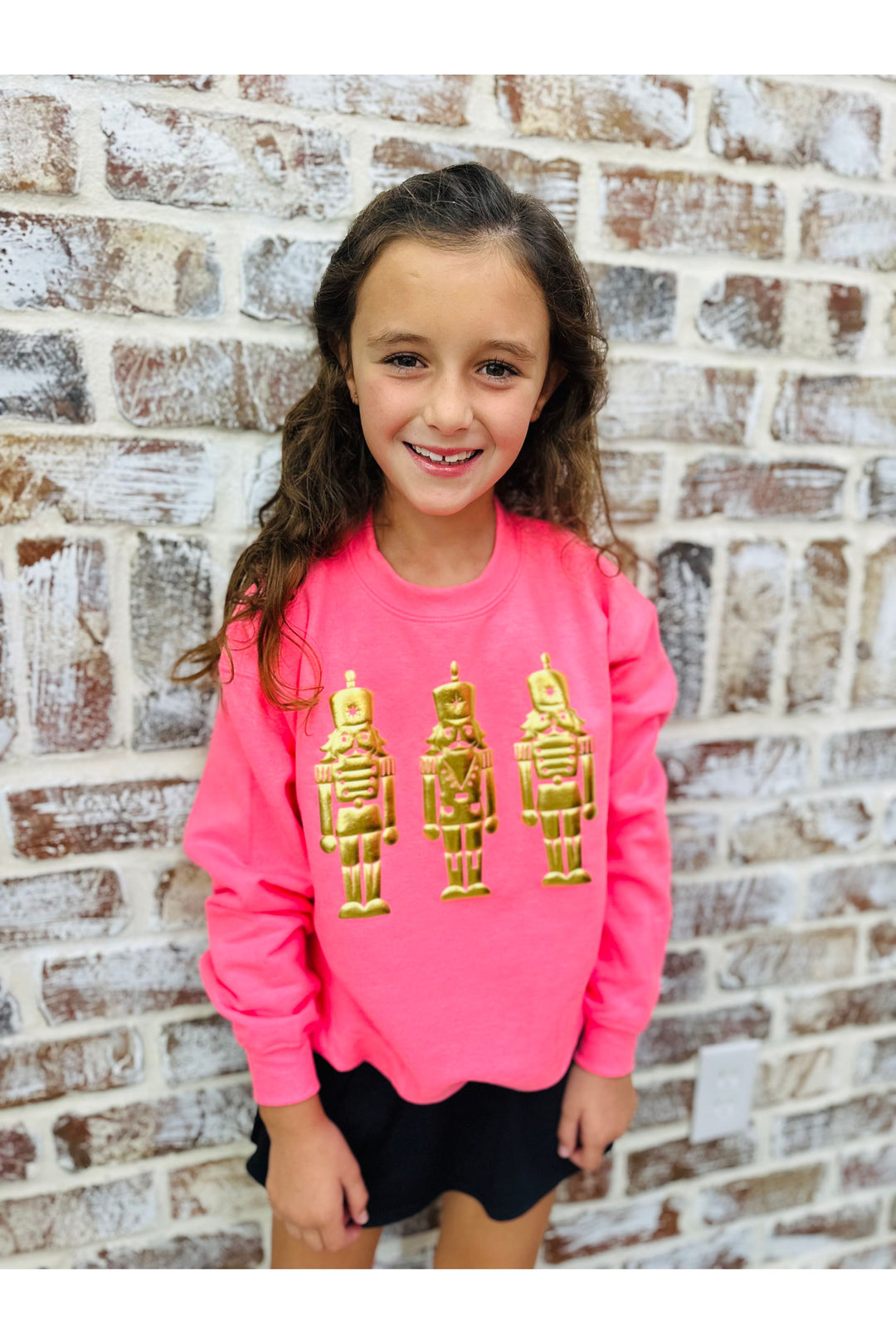 Young girl wearing a pink sweatshirt with gold nutcracker designs against a brick wall.