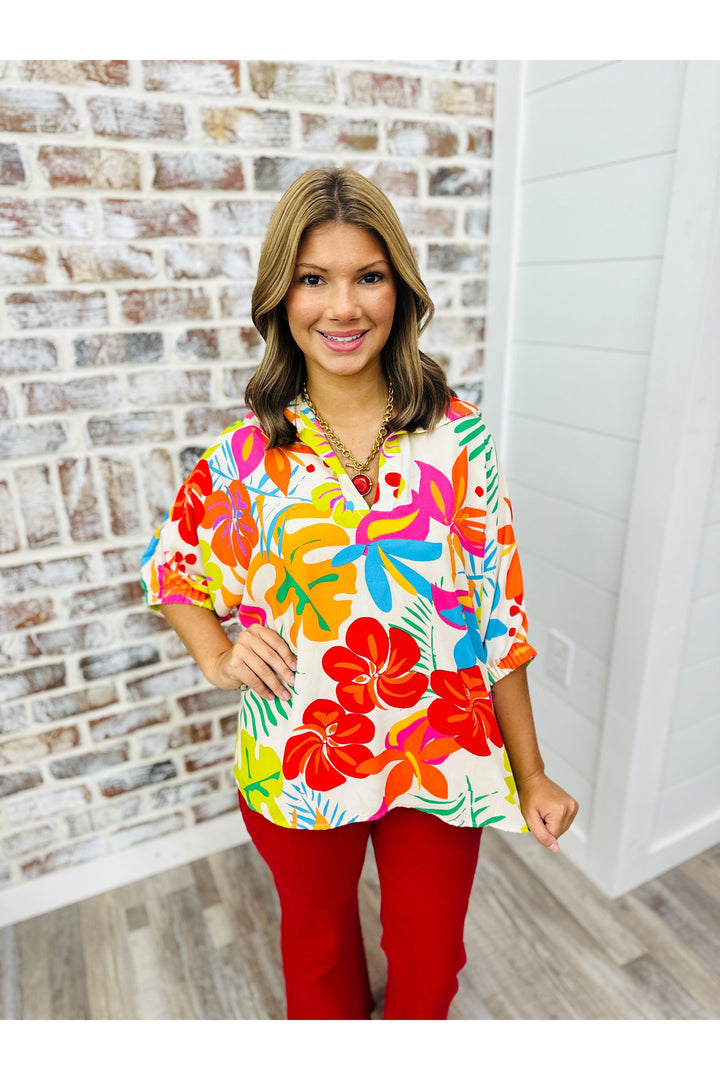 Woman wearing tropical pattern top with red pants standing against a brick wall.