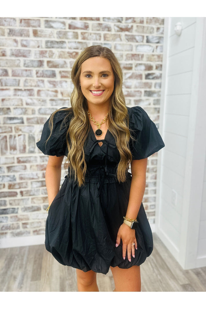 Woman wearing solid black dress with puff sleeves and bottom standing against a brick wall. Paired with Julie Vos Statement Necklace.