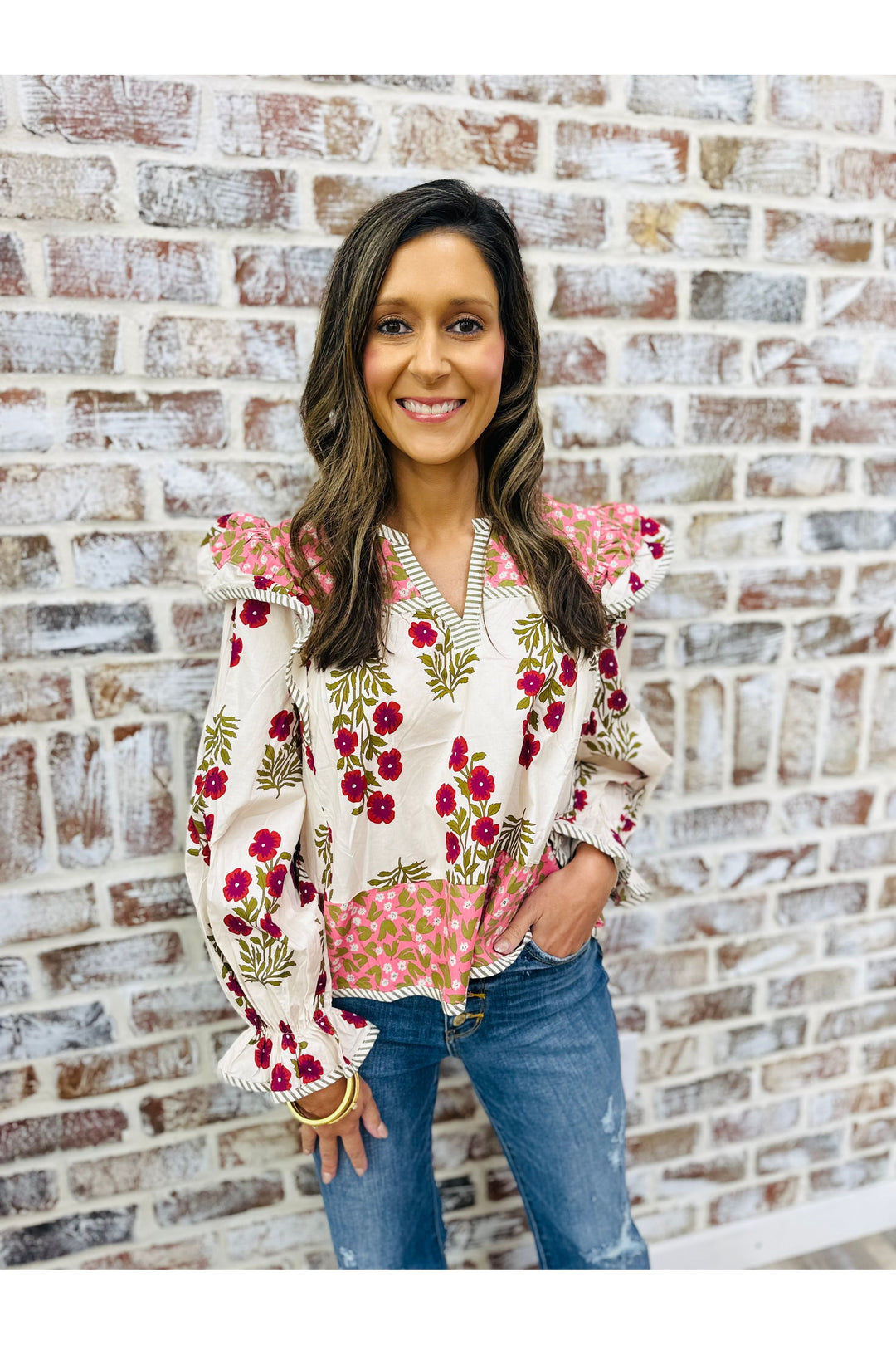 Woman wearing a floral blouse and jeans standing against a brick wall.
