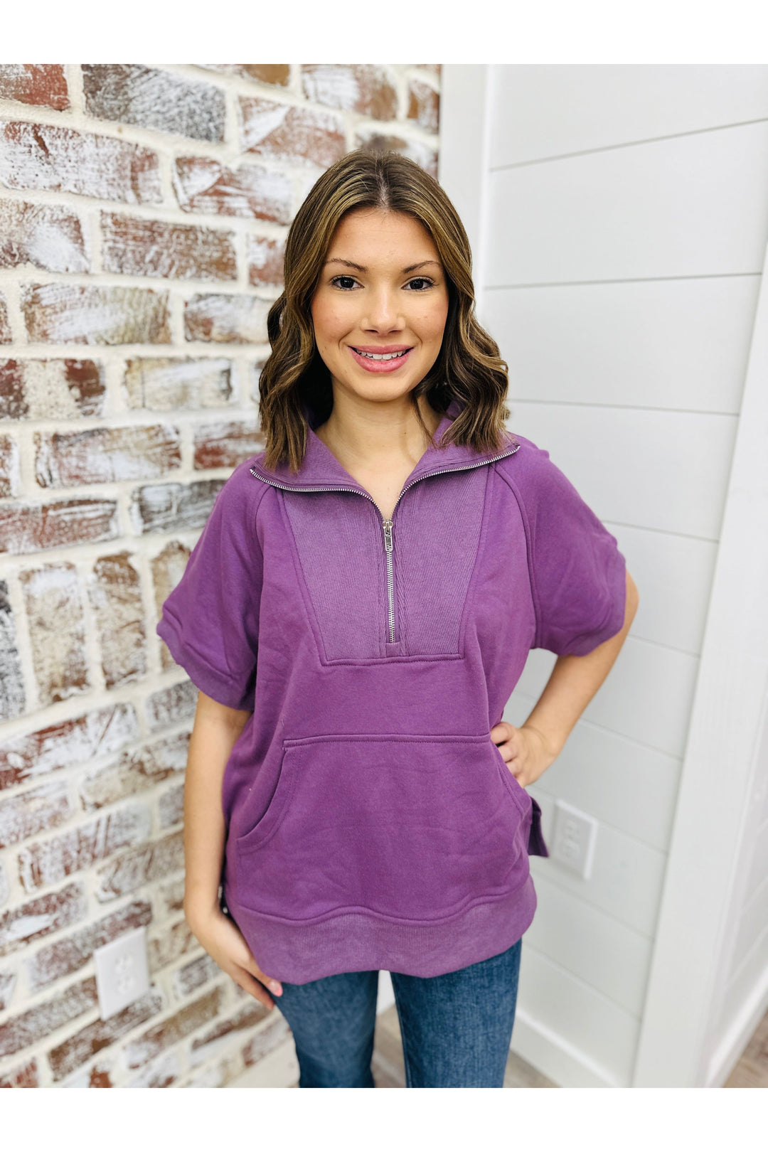 Woman wearing a plum short sleeve pullover with a zipper collar and front pocket standing against a brick wall.