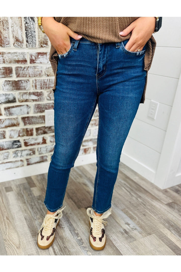 Woman wearing mid rise ankle slim straight jeans with tan and leopard sneakers standing against a brick wall. 