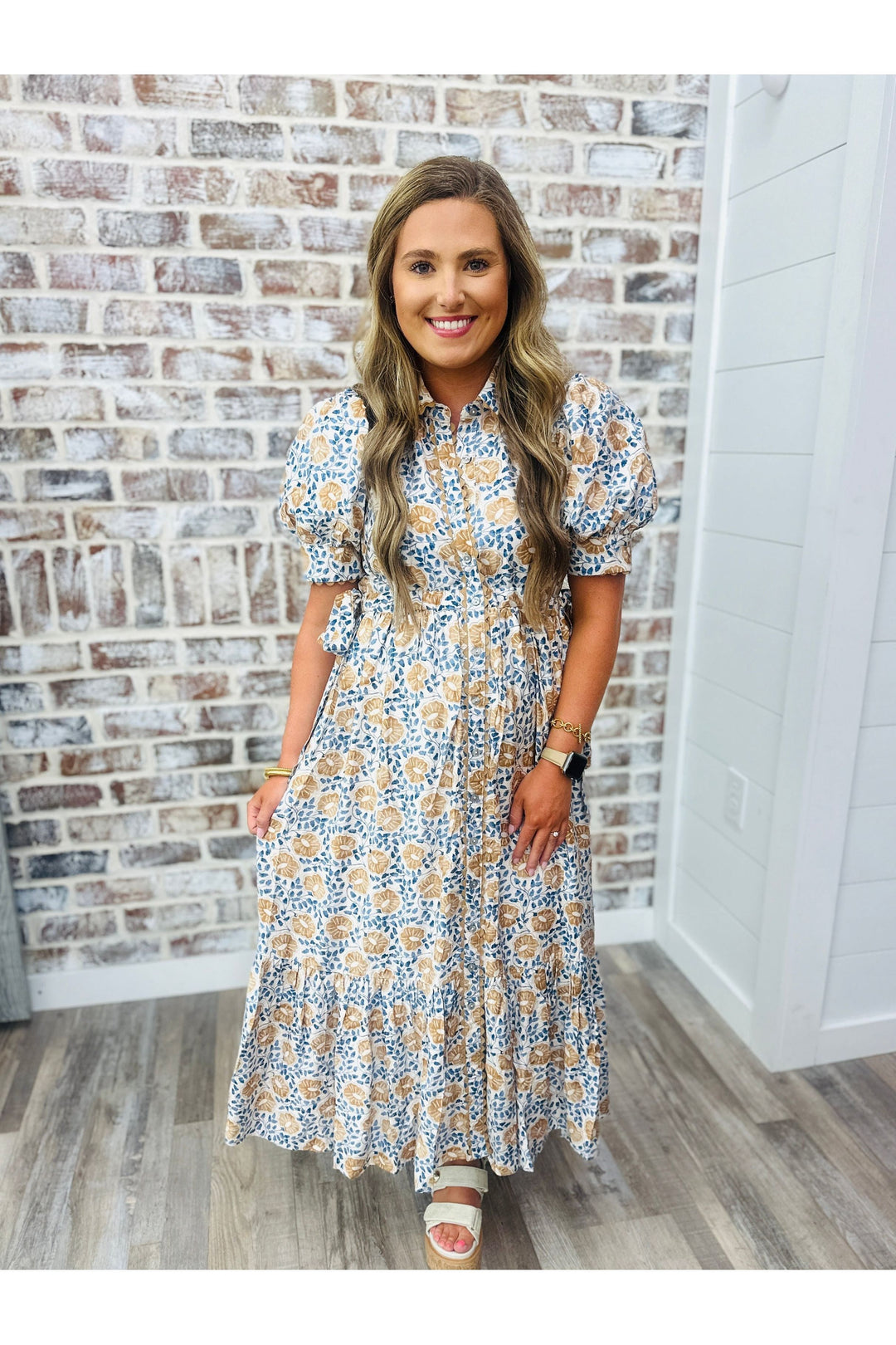 Woman wearing floral maxi dress with hues of blue and tan standing against a brick wall.