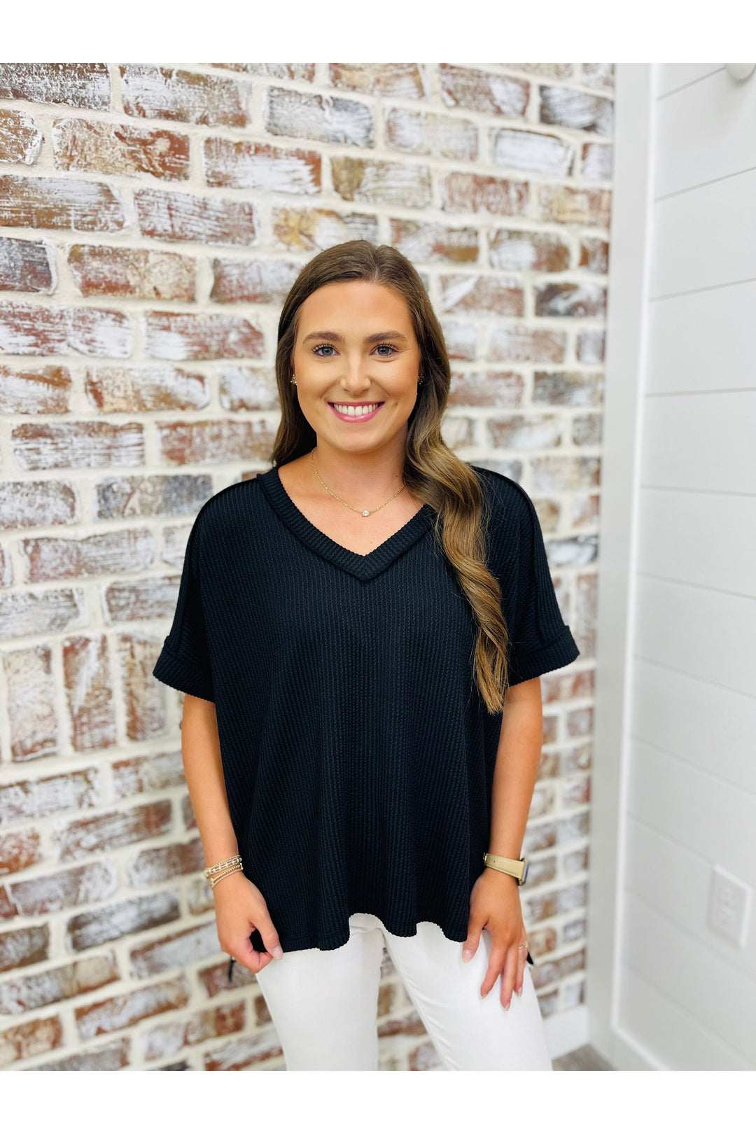 Woman wearing black waffle short sleeve top with white denim standing against a brick wall.