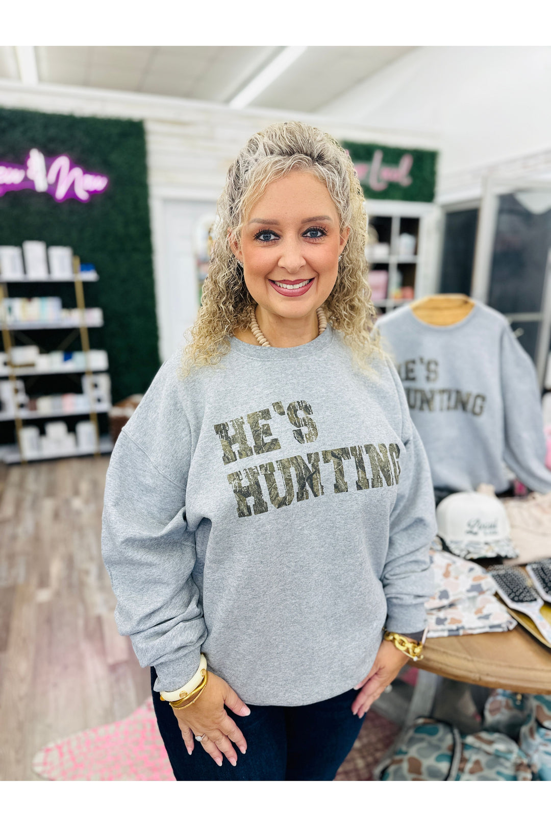 Grey sweatshirt with camo lettering saying He’s Hunting