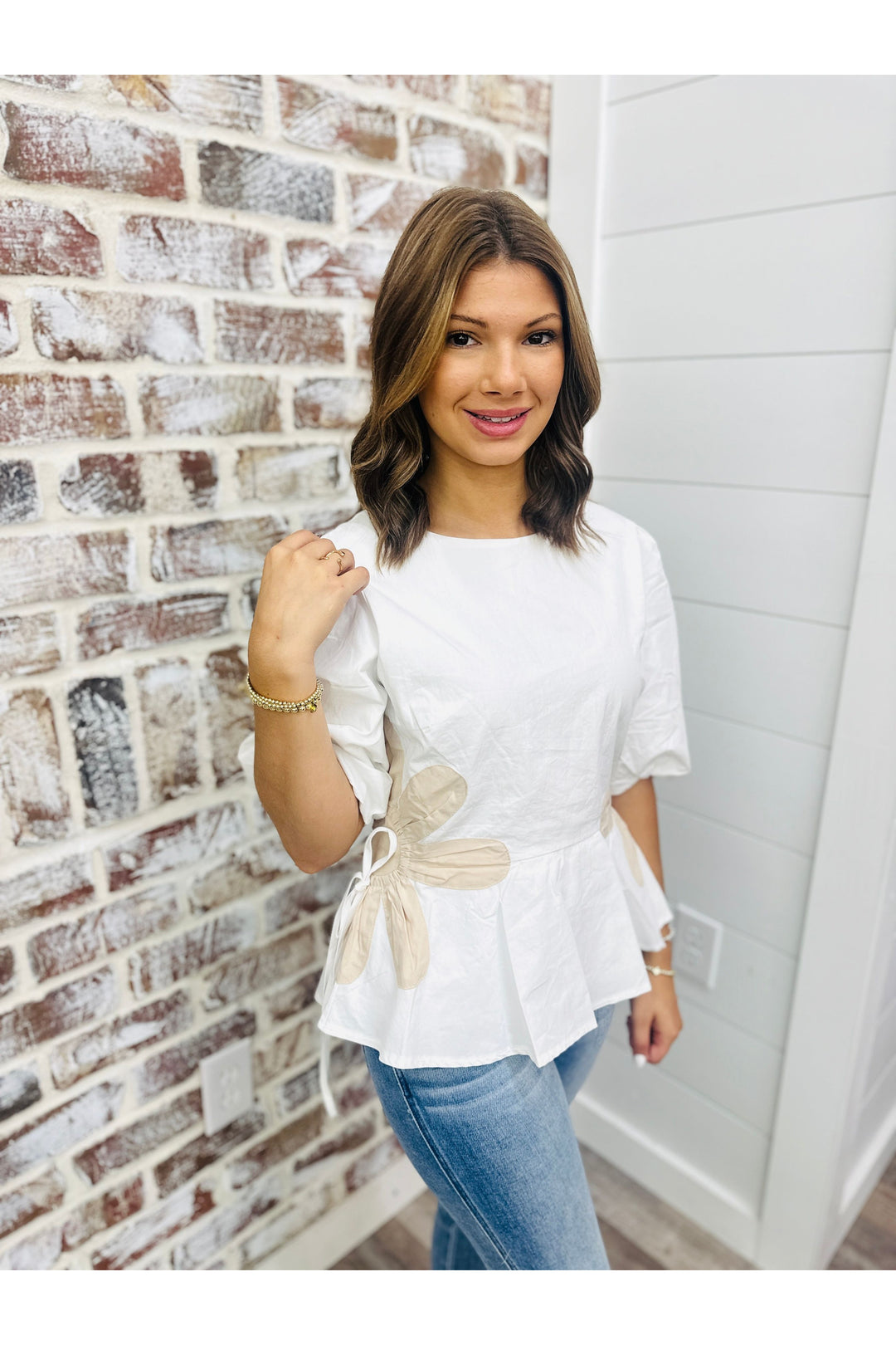 Woman wearing white ruffle top with tan flower on the side standing against a brick wall. 