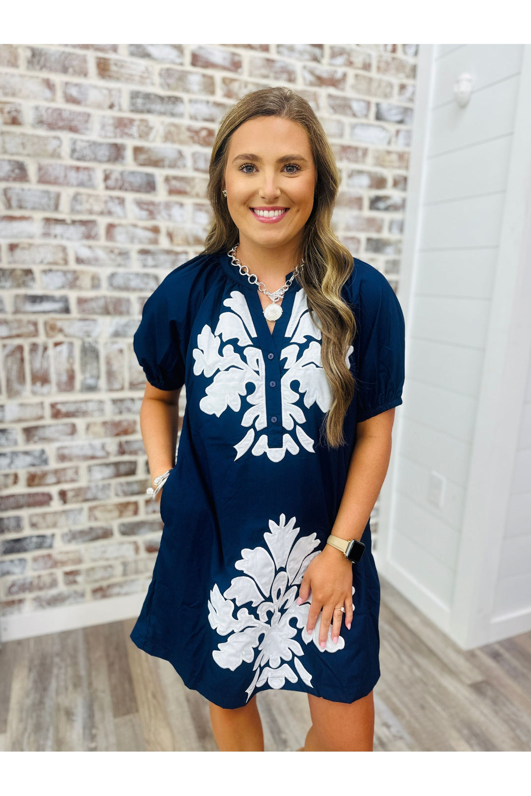 Woman wearing a navy dress with white detail standing against a brick wall.