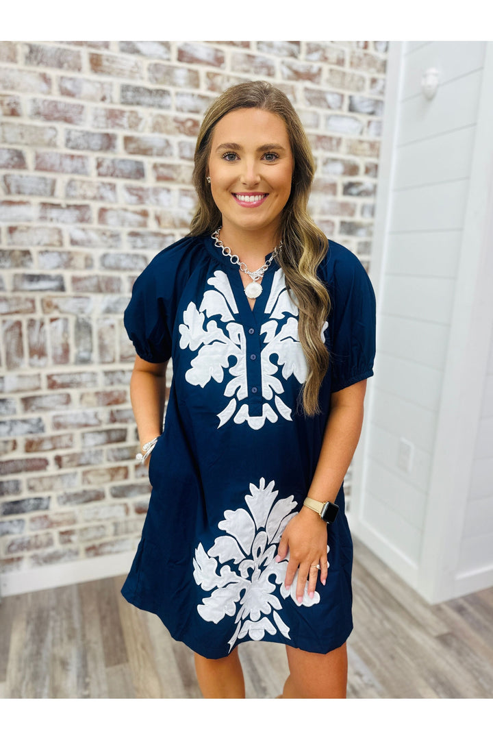 Woman wearing a navy dress with white detail standing against a brick wall.
