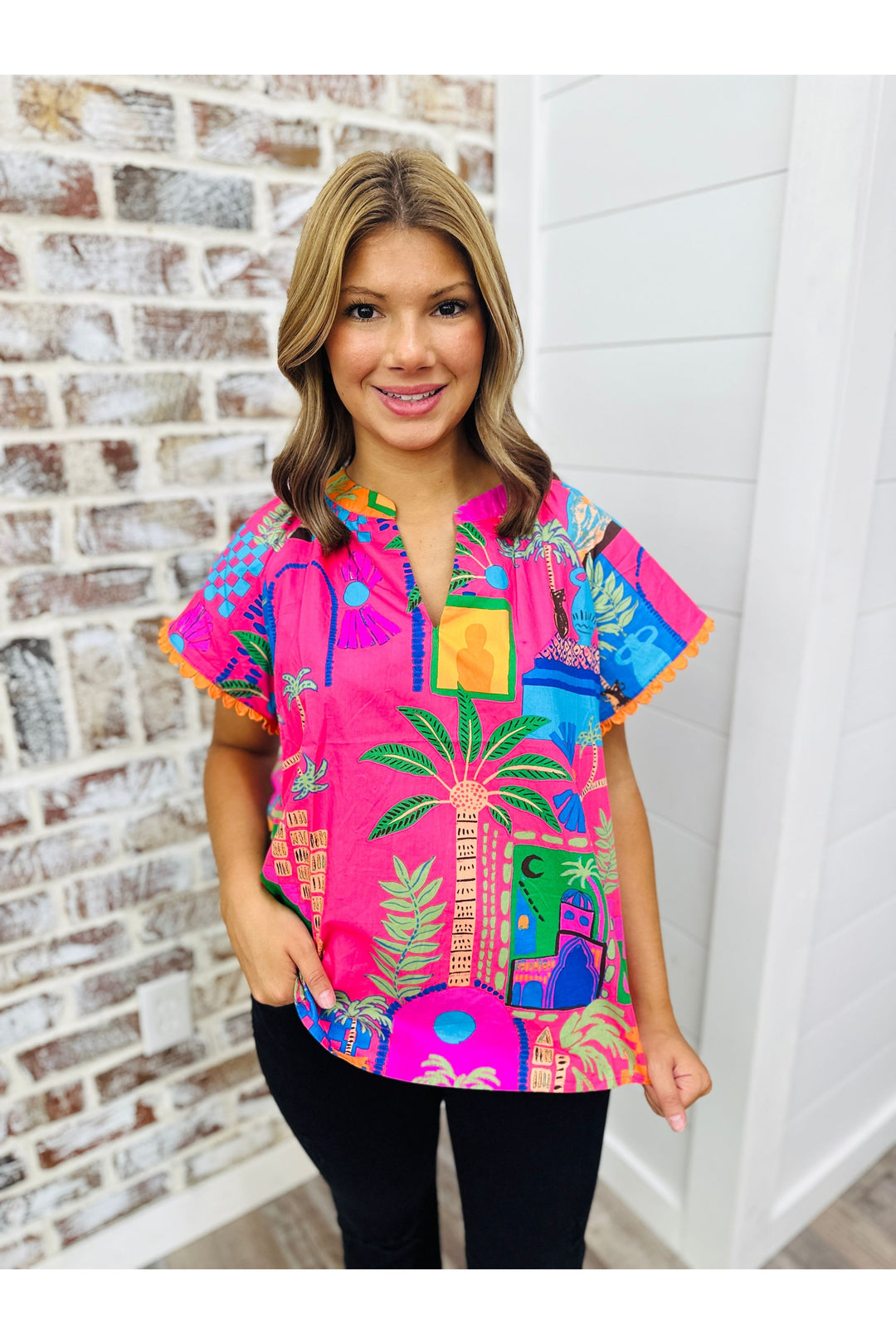 Woman wearing tropical print top with black jeans standing against a brick wall.