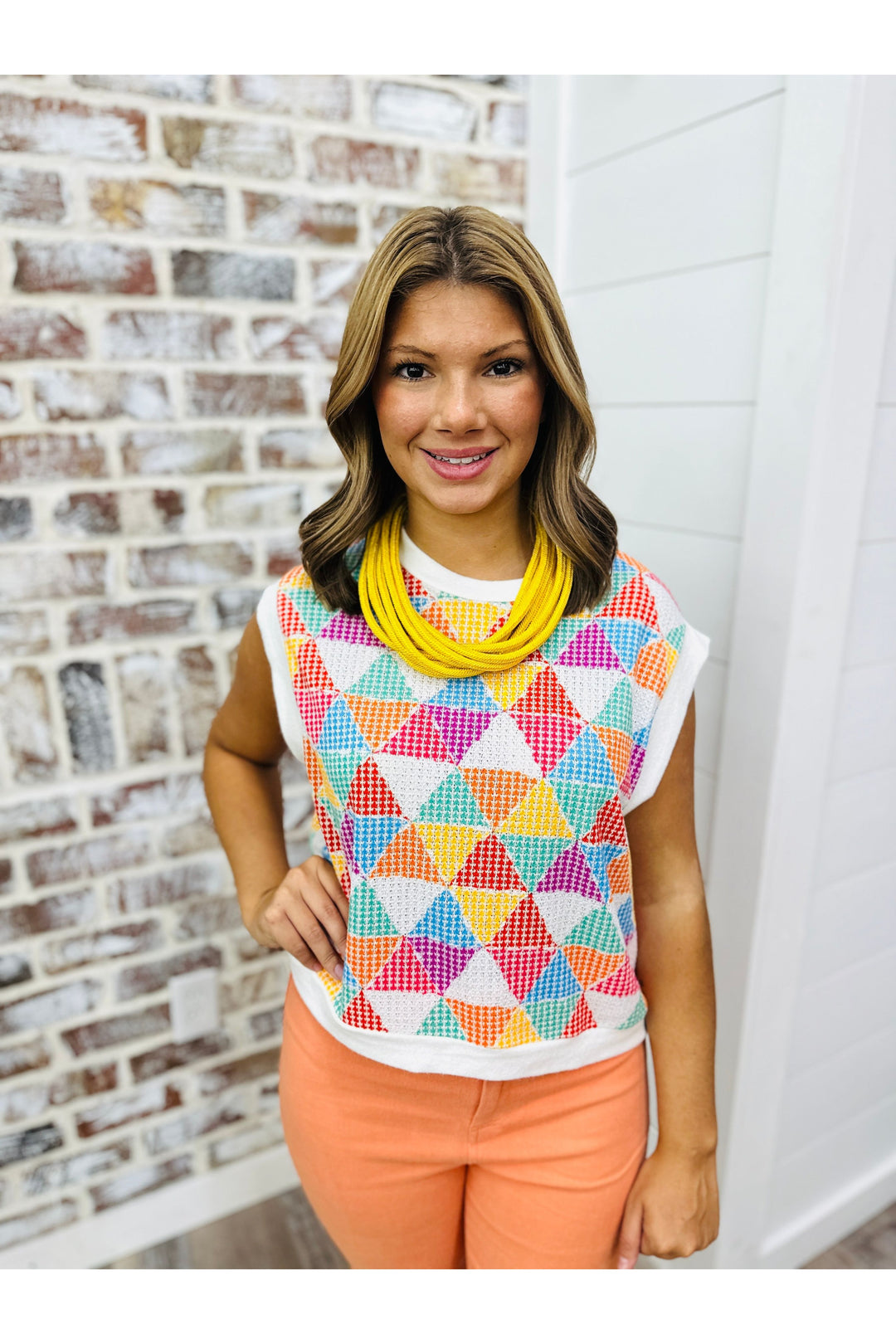 Woman wearing a colorful geometric sweater top with orange pants standing against a brick wall. Paired with a yellow strand necklace.
