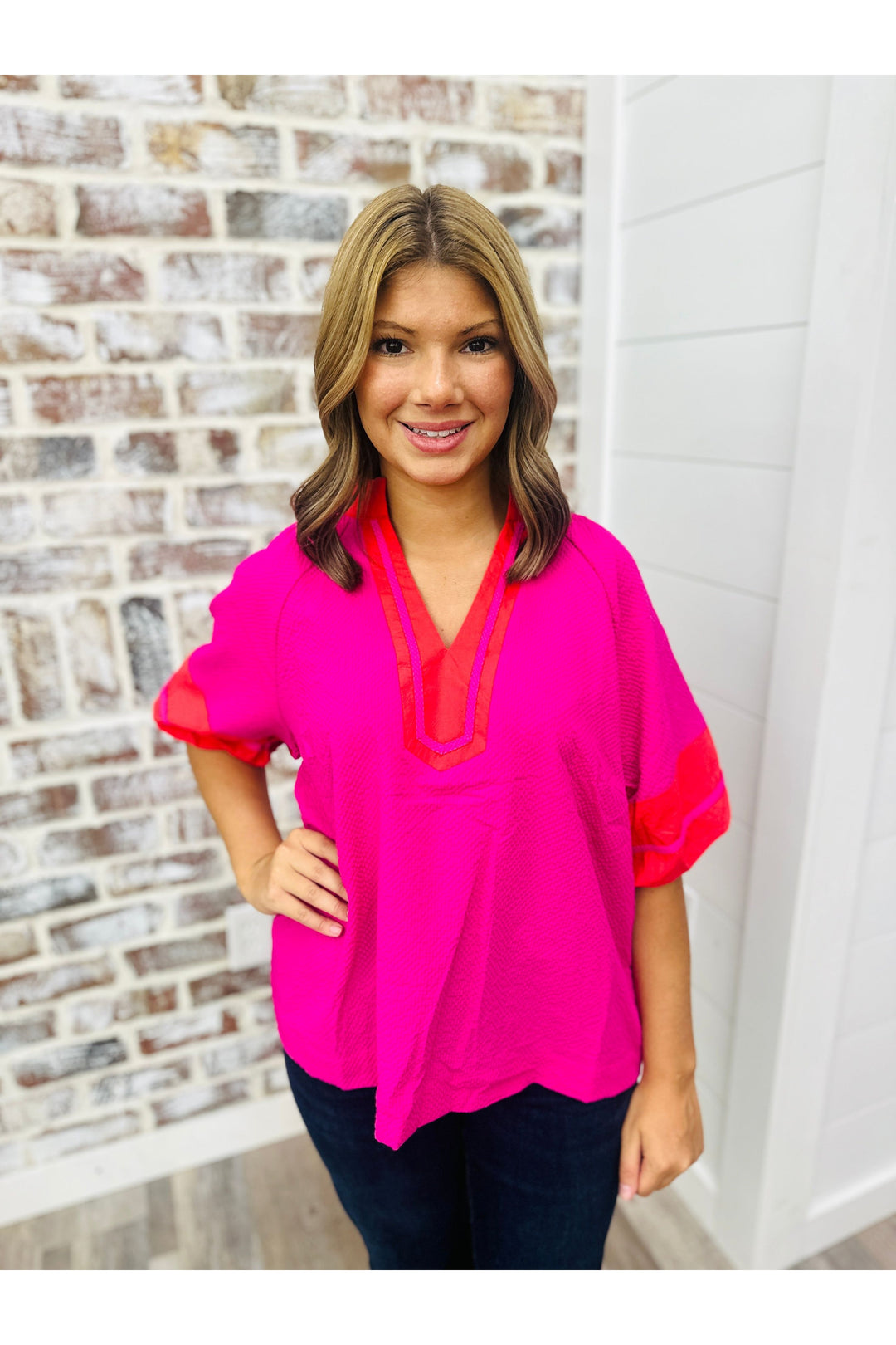 Women wearing a fuchsia pink top with red detailing with denim jeans standing against a brick wall.