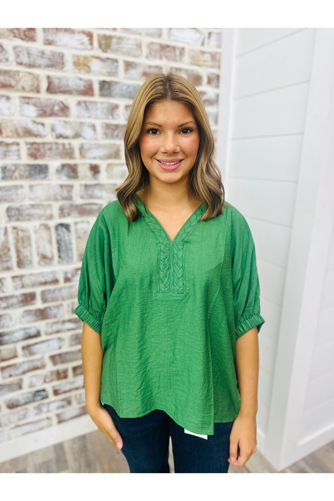 Woman wearing a fern green top with braided detail on the neckline standing against a brick wall.