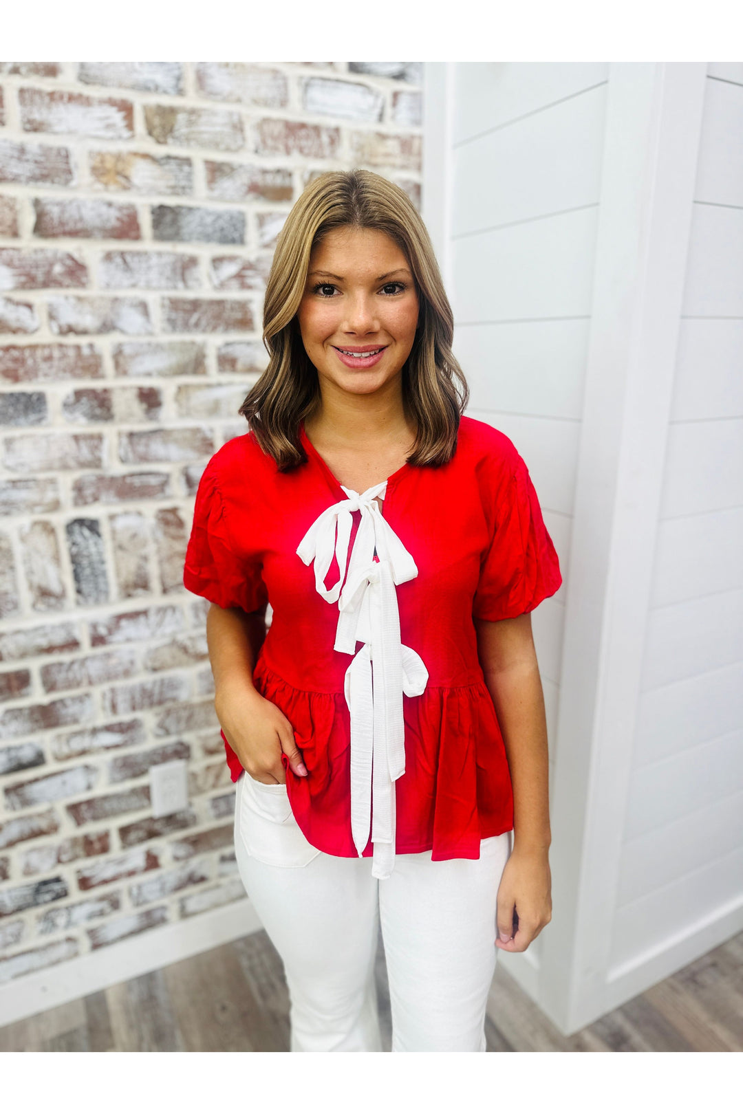 Women wearing a red top with white bows down the front with white pants standing against a brick wall.