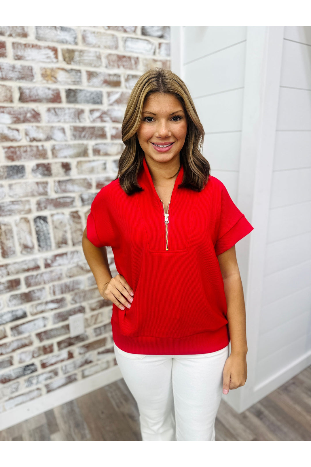 Women wearing red top with gold zipper and white pants standing against a brick wall.