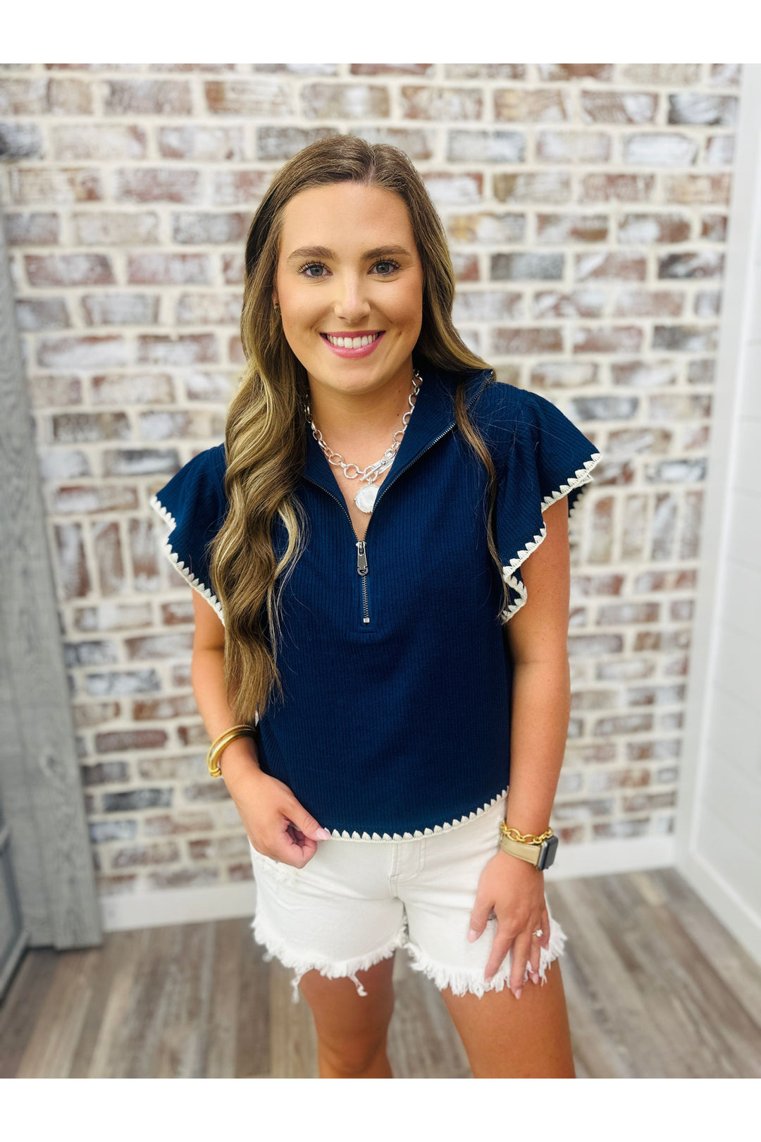 Woman wearing a navy top with zipper and embroidered trim standing against a brick wall.