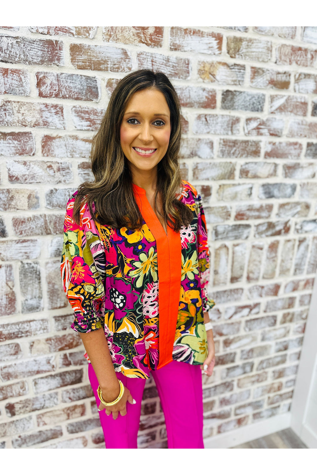 Woman wearing contrast floral print top with hot pink pants standing against a brick wall.
