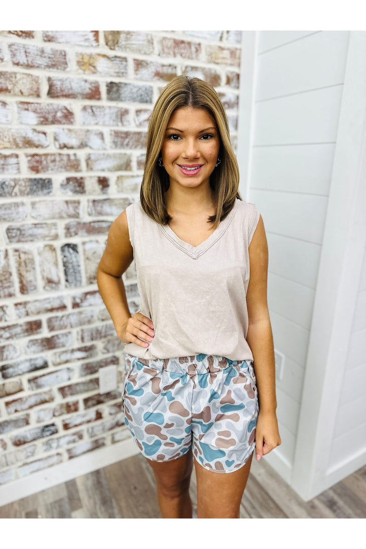 Woman wearing a acid wash ash mocha tank top with camo shorts standing against a brick wall.